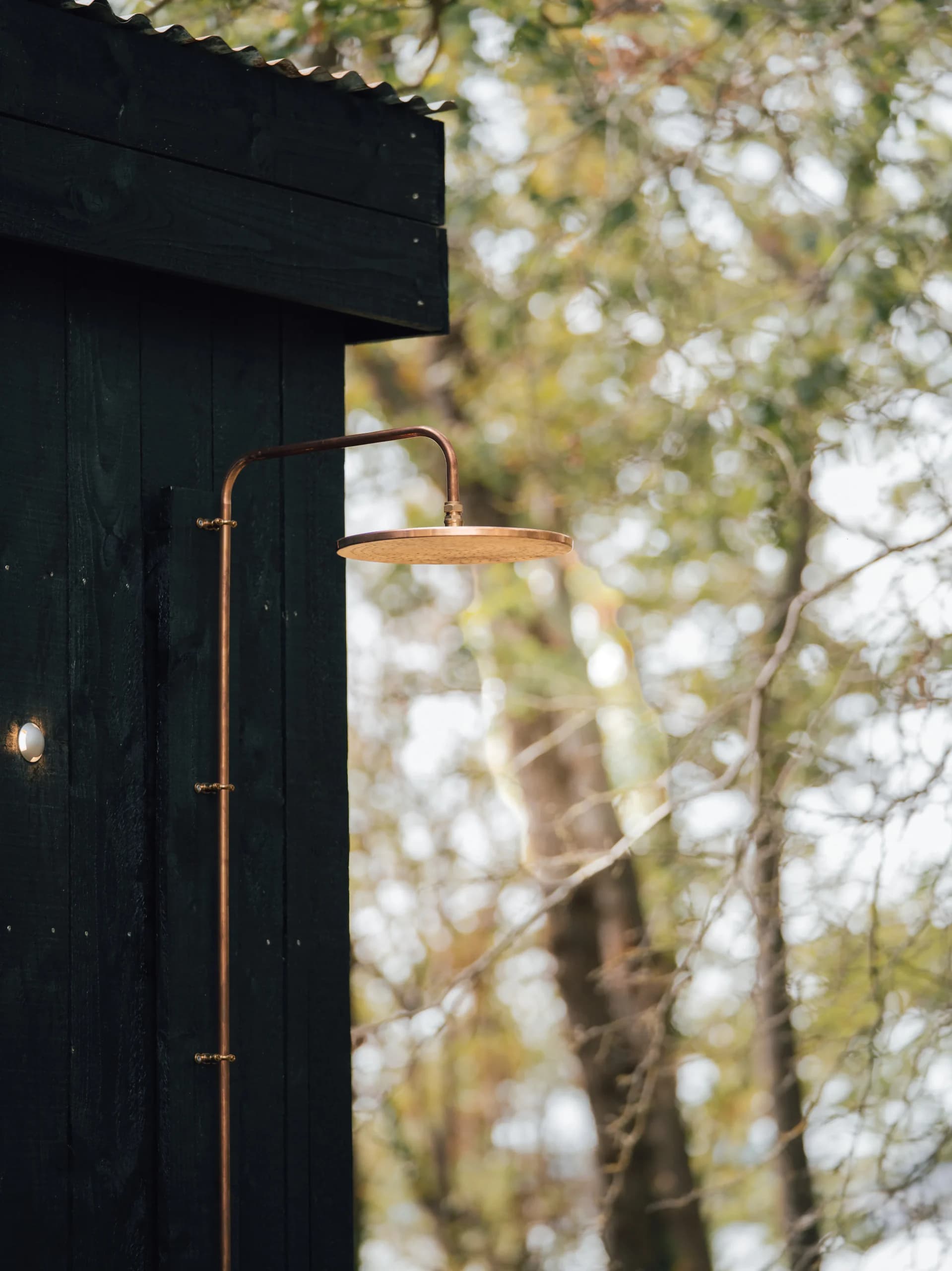 A copper shower head on the black timber wall of the sauna, with trees behind.