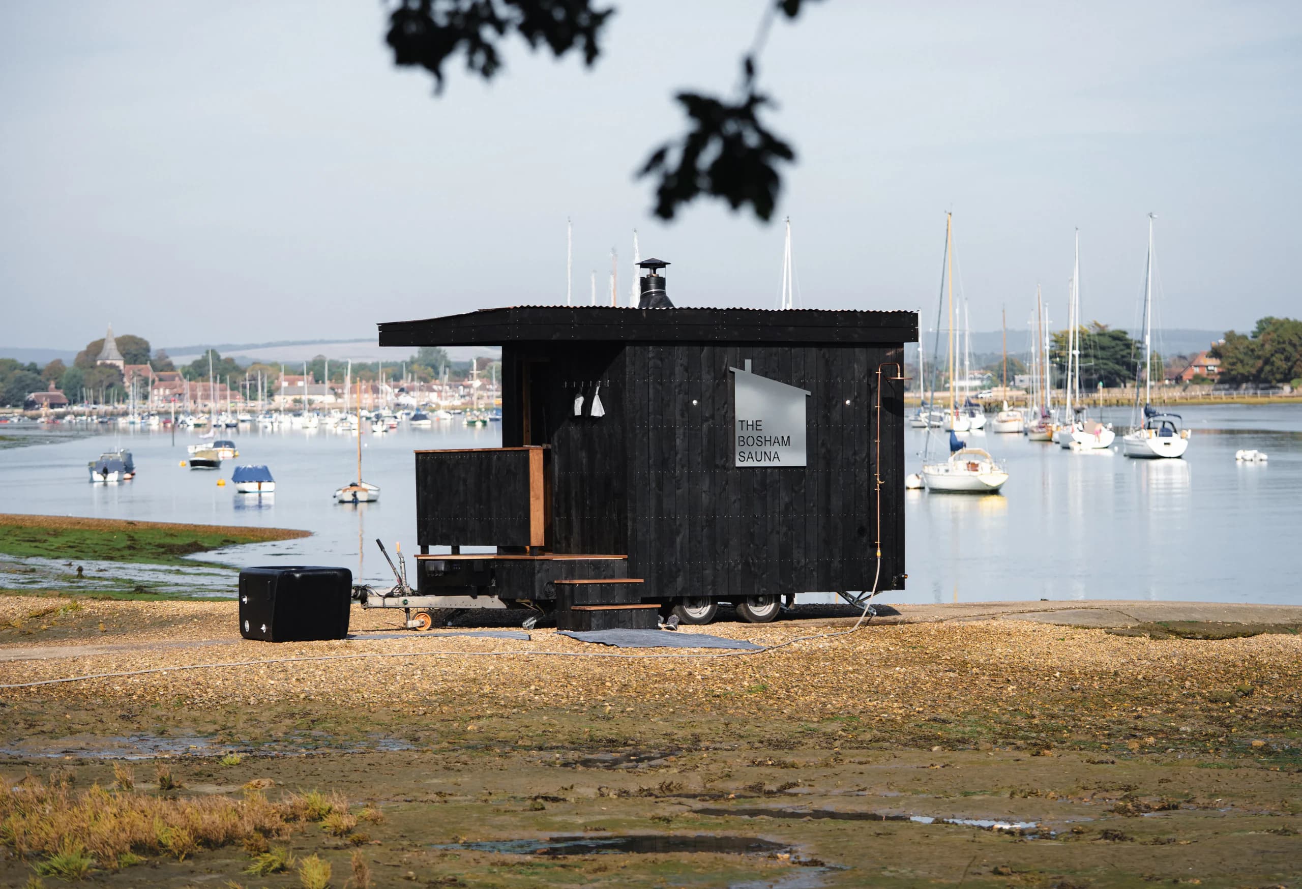 The black timber Bosham Sauna on the shore at low tide, with moored boats and the village behind.